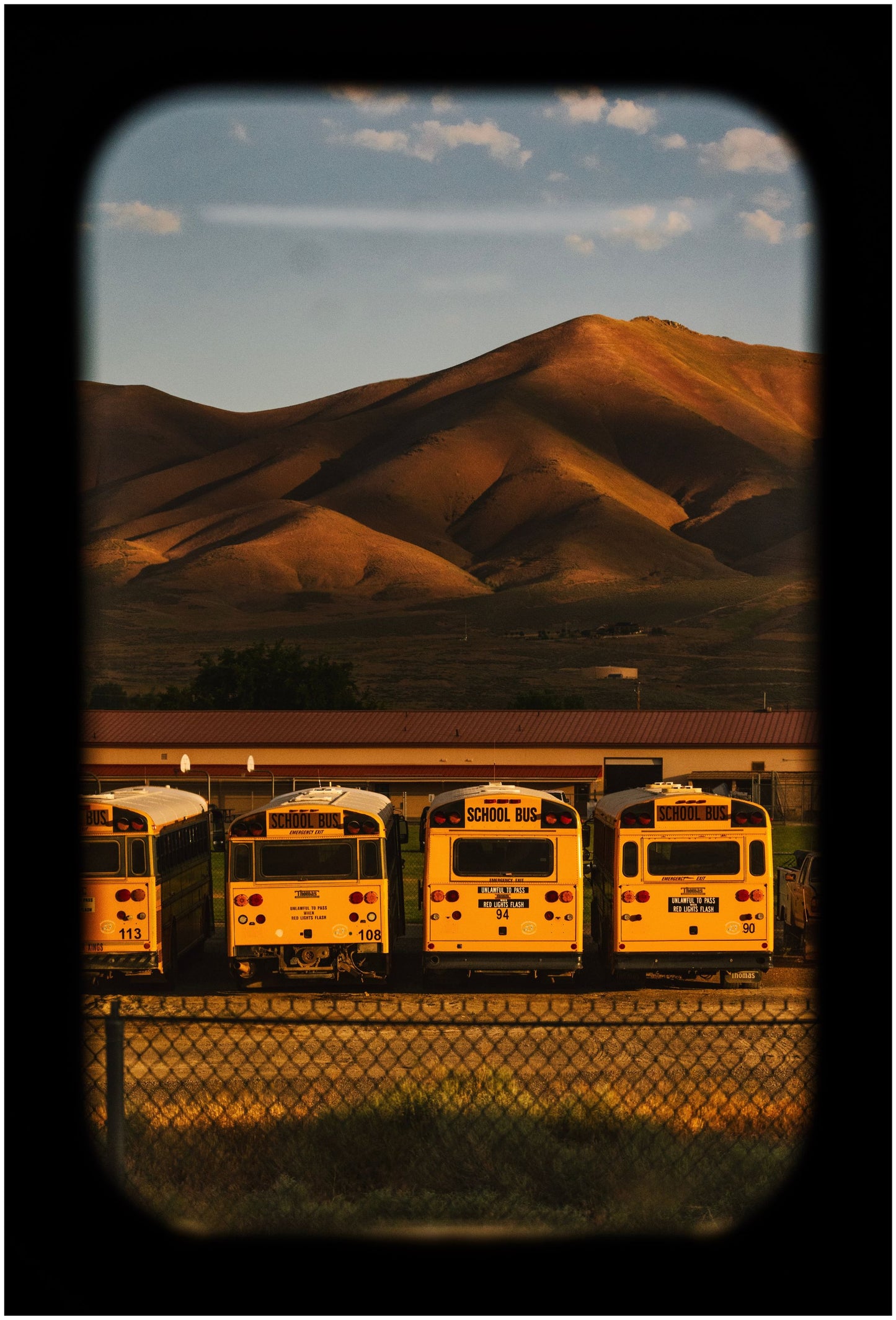 A framed artwork depicting a landscape with mountains in the background and several yellow school busses in the foreground, captured through what appears to be a window with a dark frame.