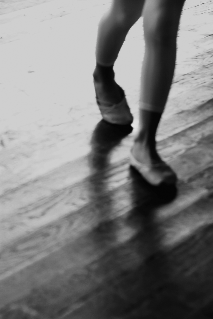 A black and white image of a person's lower legs and feet in motion, suggesting a dance movement, on a wooden floor.