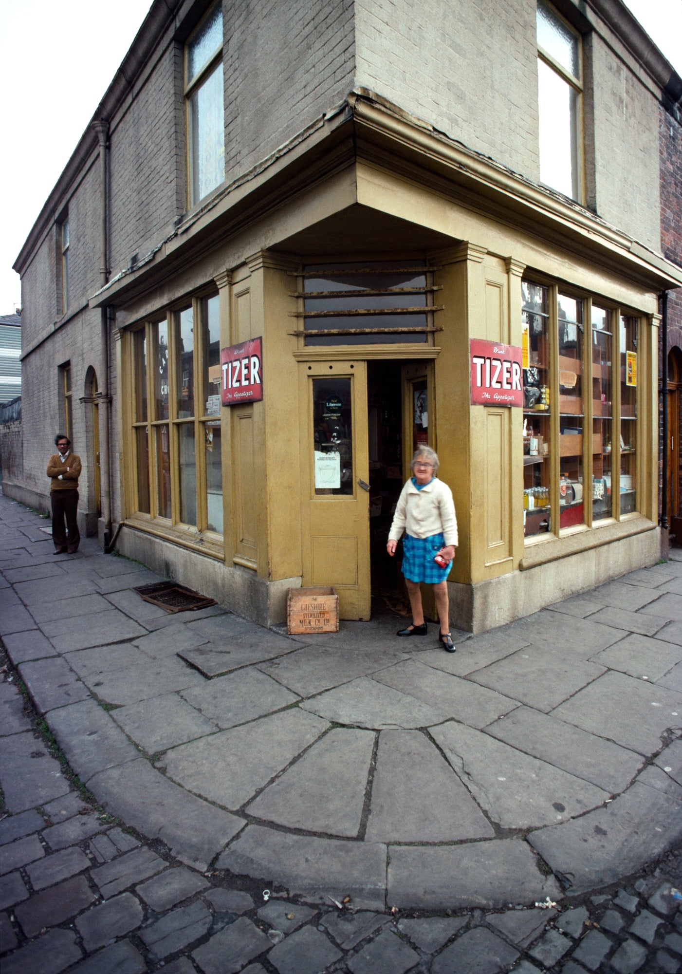 A photograph of a lady standing in the doorway of a shop with yellow walls, with a man standing around the corner