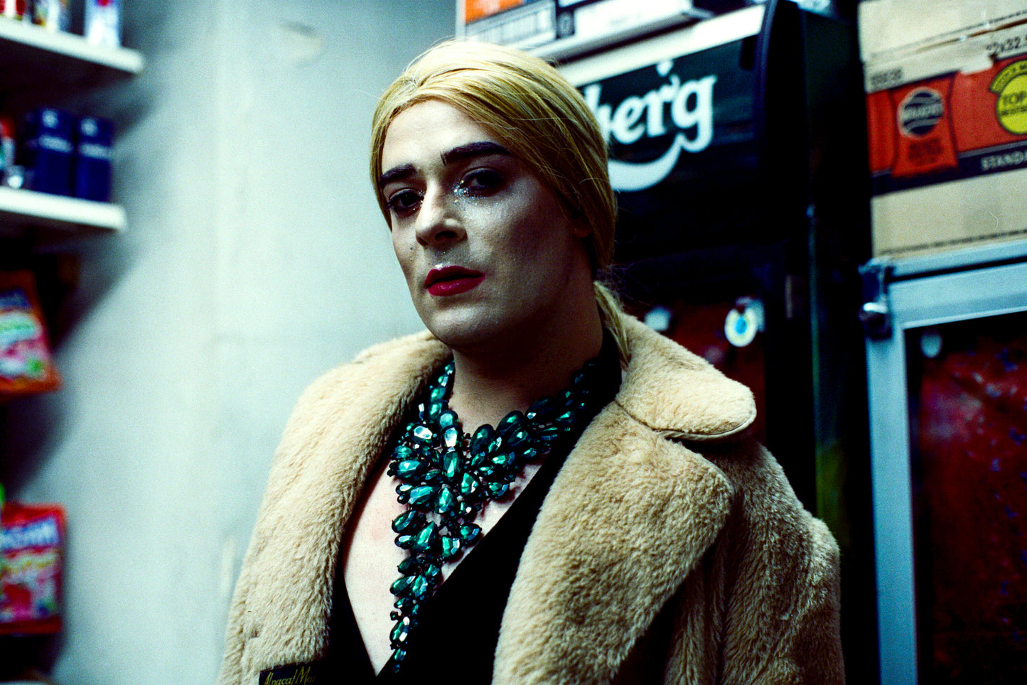 A photograph of a person with blond hair, red lipstick and a green necklace and a fur coat.