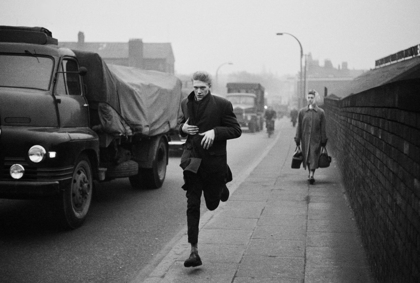 Black and white photograph of a man running on a street with vehicles in the background, including a truck on the left and a car further down the road.