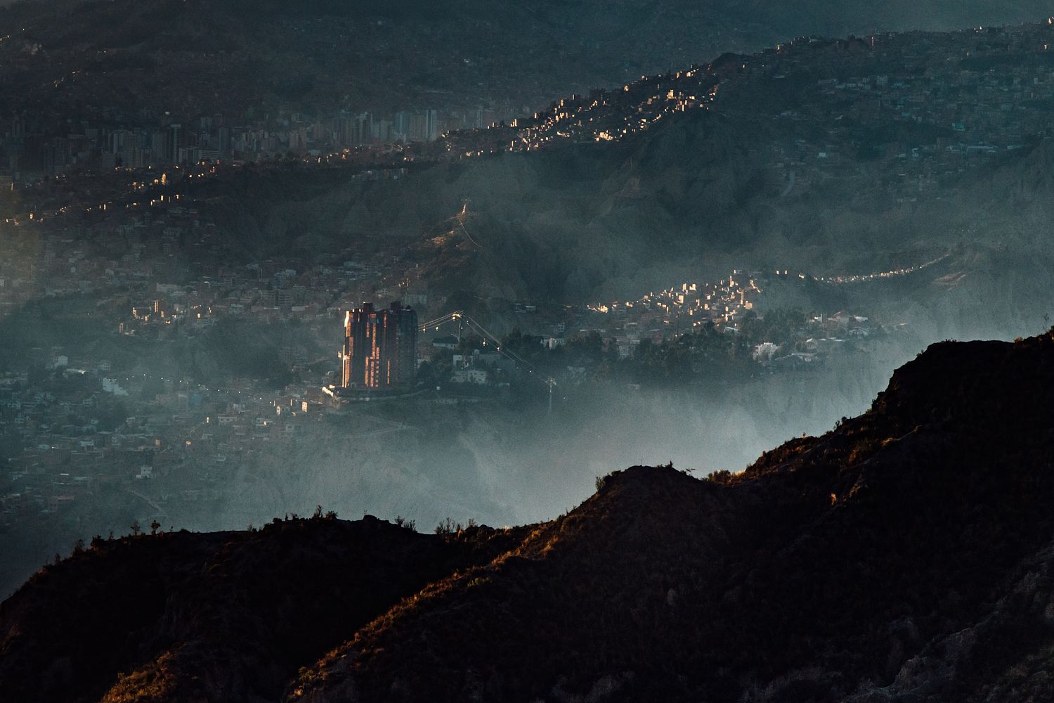 An image of a city in the mountains taken from a viewpoint. With fog in the foreground and a towering city block as the main focus of the image. 
