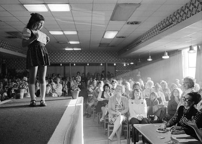 Black and white photo of a person on a stage with a crowd in the background, likely a part of a carnival queen competition.