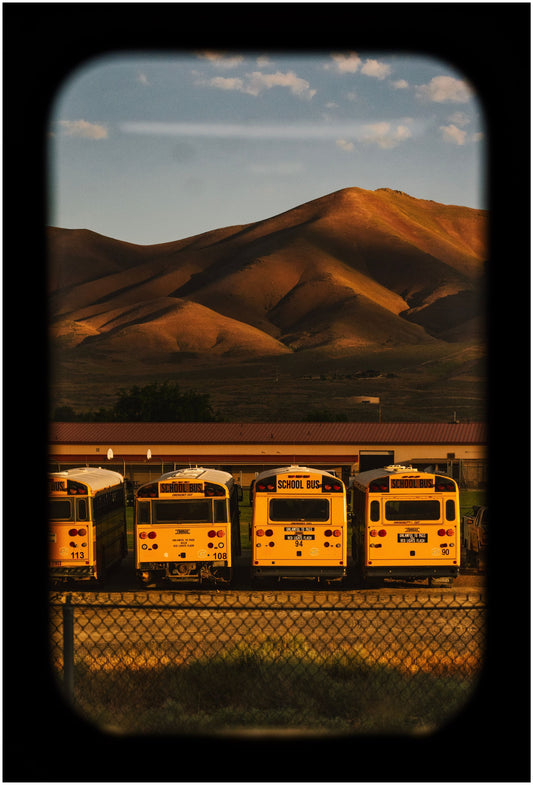 A framed artwork depicting a landscape with mountains in the background and several yellow school busses in the foreground, captured through what appears to be a window with a dark frame.