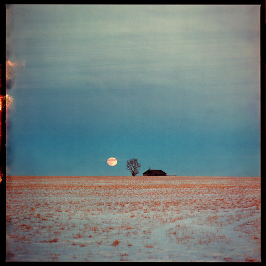 An analogue picture of a landscape with a barn in the far distance with a tree next to it and the moon behind it