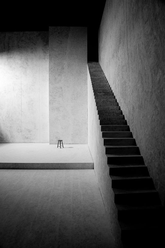A black and white photograph of a staircase leading to a dark area, with a chair visible in the distance.