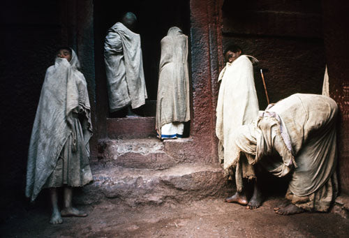 A black and white photograph featuring a group of Ethiopian Orthodox monks in traditional attire, standing near a historic site.