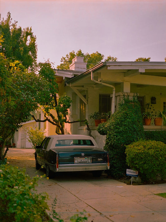 A fine art print featuring a classic car parked in front of a house with a warm-toned sunset in the background, displayed with a certificate of authenticity.