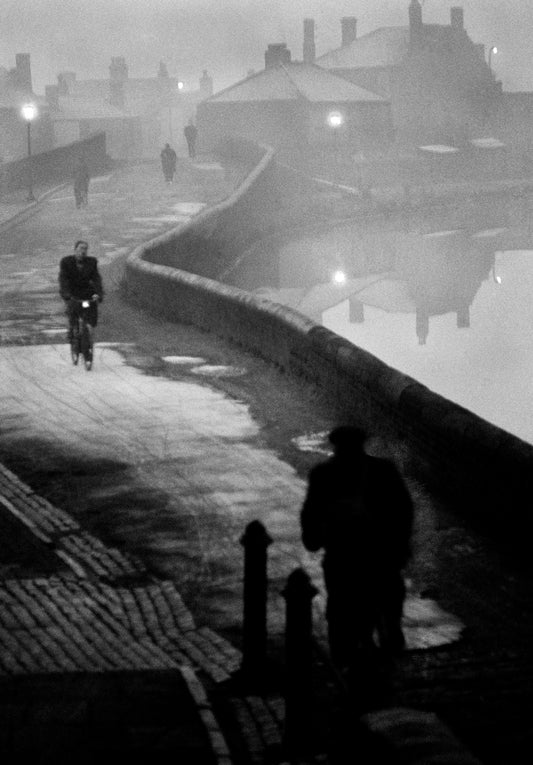 A black and white photo of a quiet street at dusk with a few people and a bicyclist, reflecting the artistic style of John Bulmer.