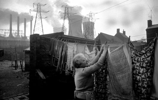 Black and white photo of a woman hanging laundry out to dry, with a power station in the background.