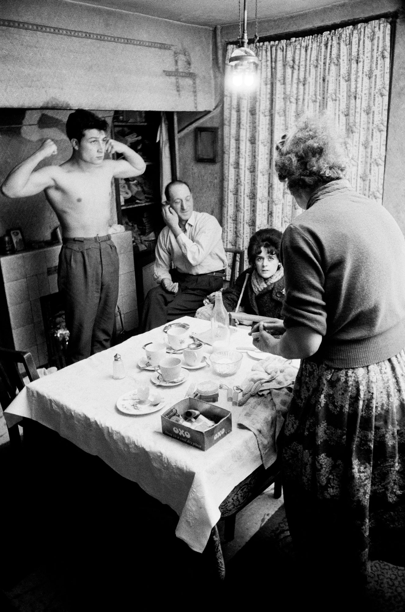 Black and white photograph of a group of people seated around a table, with a man displaying a muscular pose in the background.