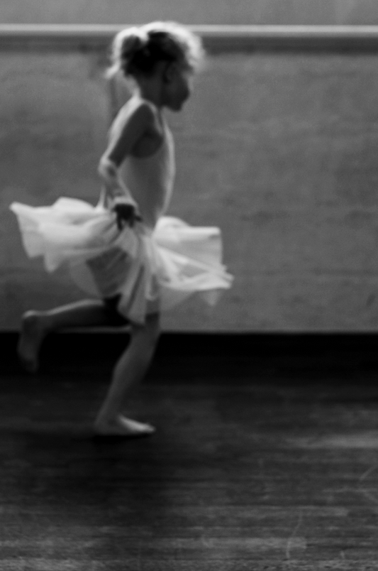 Black and white image of a person dancing, likely a child, in a room with a plain background.
