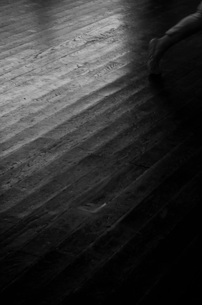 A black and white image of a silhouette of a person dancing on a wooden floor, with the shadow of a dancer cast on the floor.