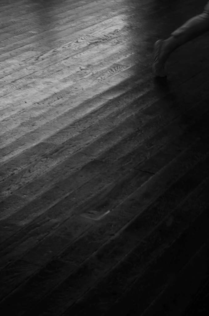 A black and white image of a silhouette of a person dancing on a wooden floor, with the shadow of a dancer cast on the floor.
