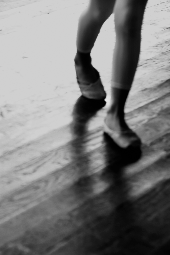 A black and white image of a person's lower legs and feet in motion, suggesting a dance movement, on a wooden floor.