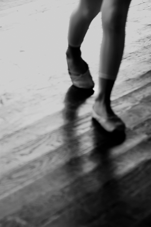 A black and white image of a person's lower legs and feet in motion, suggesting a dance movement, on a wooden floor.