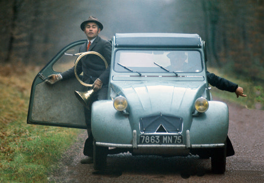 A photograph of Two men in a car one s carrying a musical instrument while the other drives.