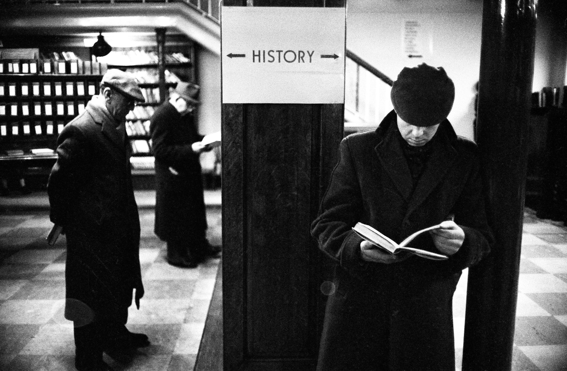 Black and white photograph of people reading in a library setting, with a sign above them that says 'HISTORY'.