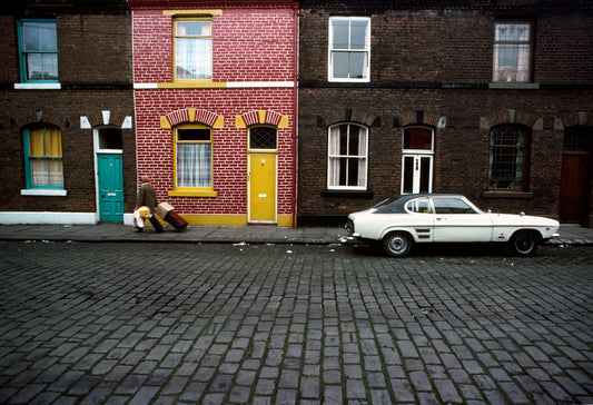 A photograph featuring a white Ford Capri parked on the side of a street with red and yellow houses in the background, and a person standing on the sidewalk with a suitcase.