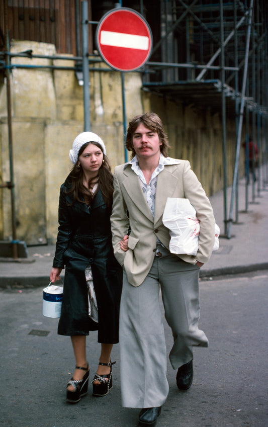 A photograph of a man and a woman walking down the street, with the man wearing baggy trousers and a flannel shirt, the woman wearing a headscarf.