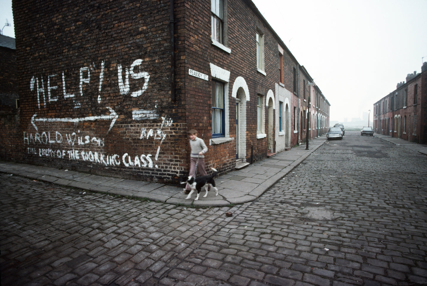 A photo of a street scene with a piece of graffiti on a wall saying 'HELP US THE WORKING CLASS!' There is a child walking their dog on the sidewalk.