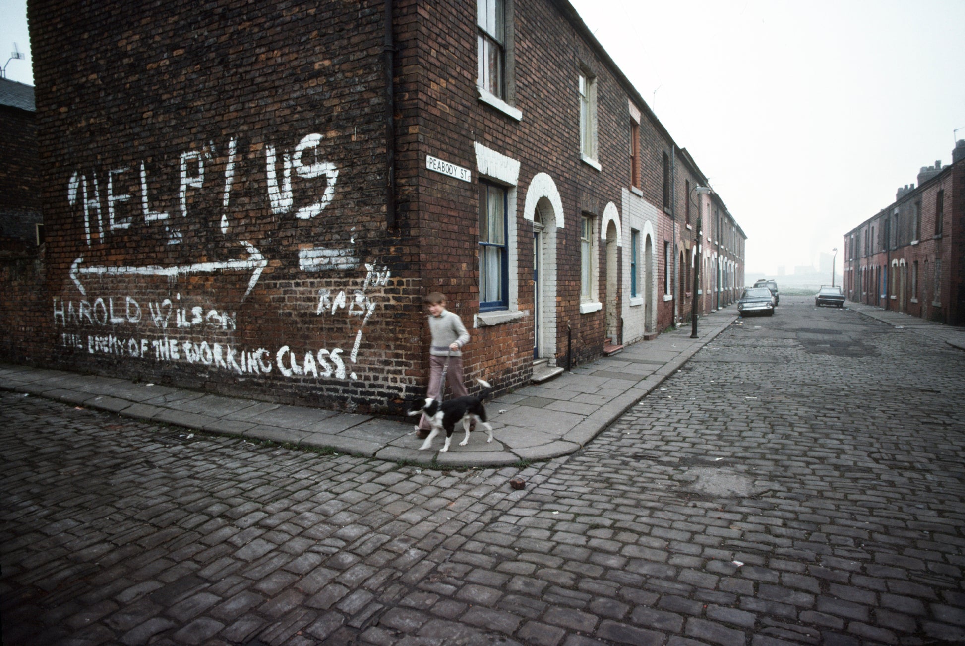 A photo of a street scene with a piece of graffiti on a wall saying 'HELP US THE WORKING CLASS!' There is a child walking their dog on the sidewalk.