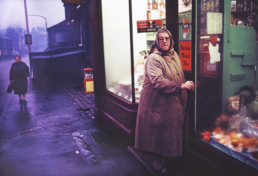 A photograph of an elderly woman standing outside a shop, looking towards the camera.
