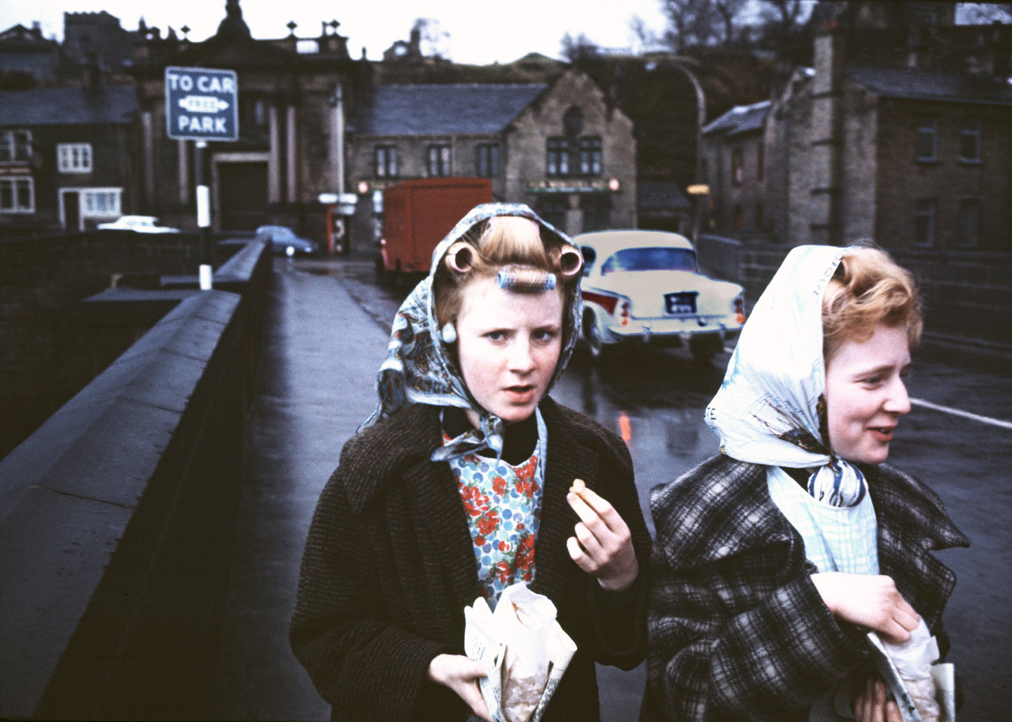 A photograph featuring two women on a sidewalk, with a car and a truck in the background, indicating a vintage 1960s setting.