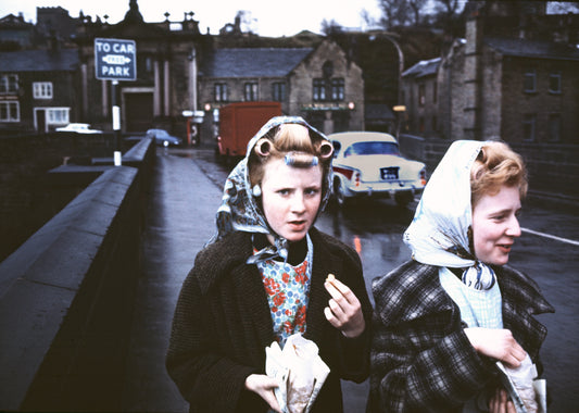 A photograph featuring two women on a sidewalk, with a car and a truck in the background, indicating a vintage 1960s setting.