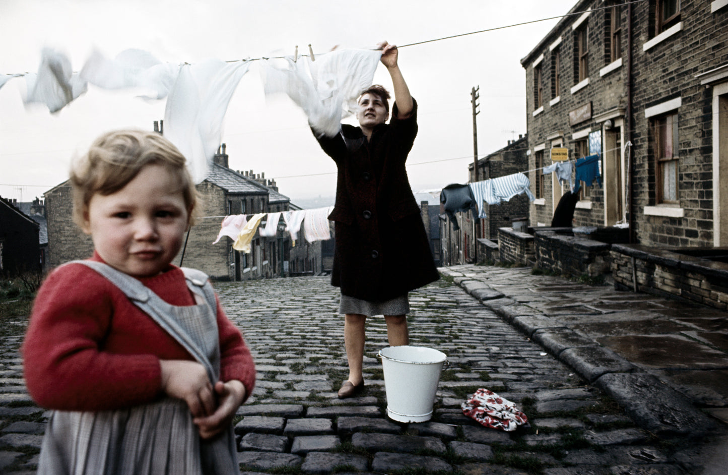 A photograph featuring two individuals, one hanging laundry on a line and the child looking on, with a backdrop of terraced houses and a gray sky.