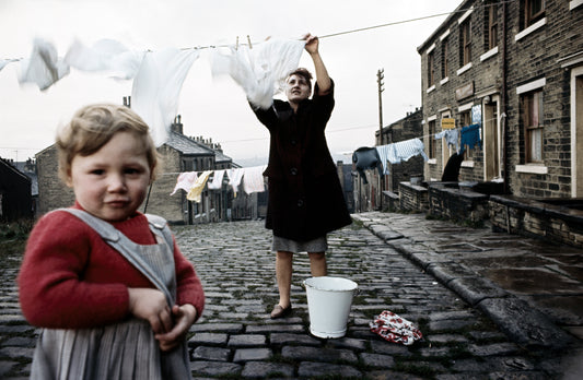 A photograph featuring two individuals, one hanging laundry on a line and the child looking on, with a backdrop of terraced houses and a gray sky.