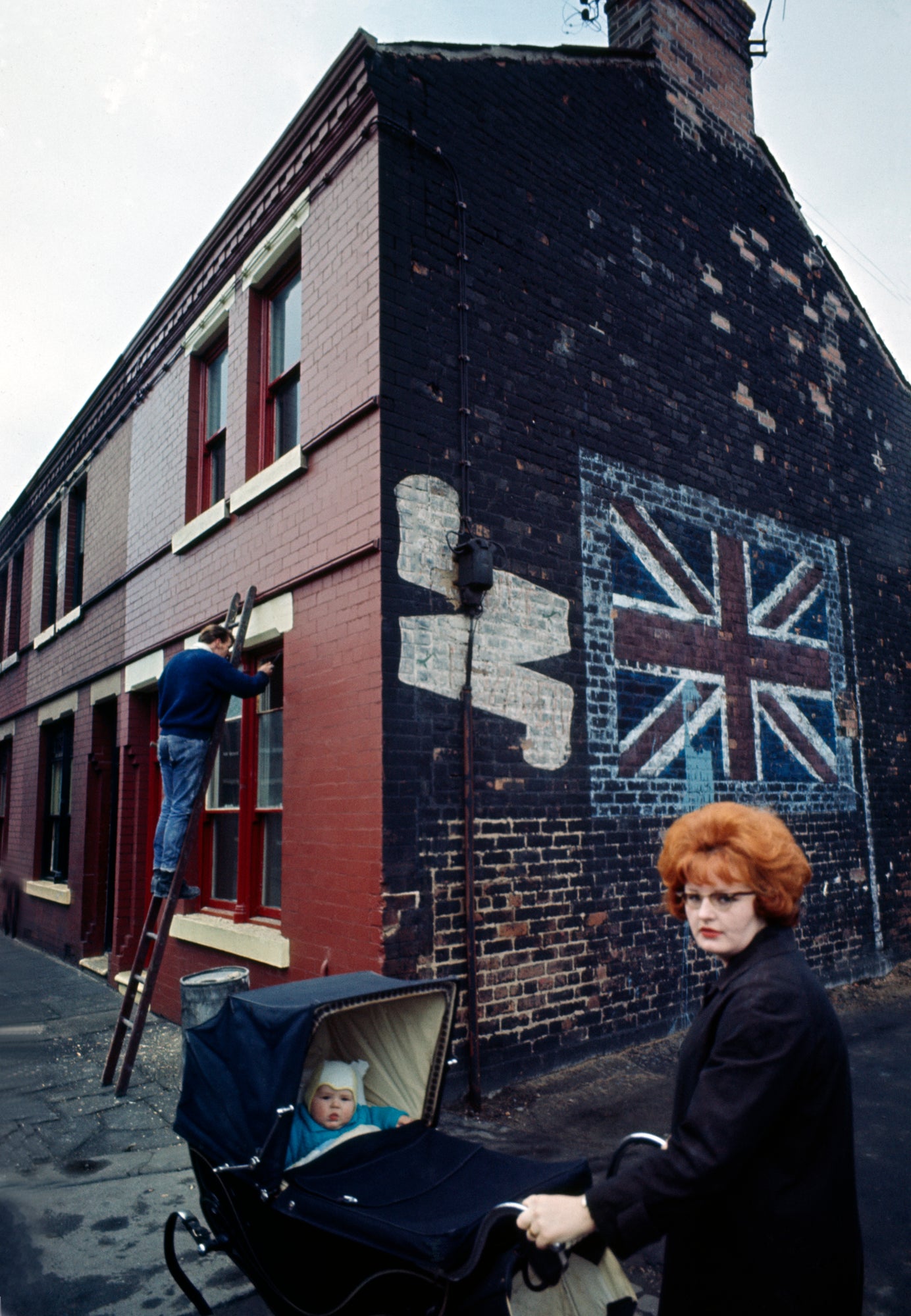 A photograph featuring a part of a building with a 'Union Jack' flag design on its side, with a man working on the building's facade and a woman holding with a baby stroller.