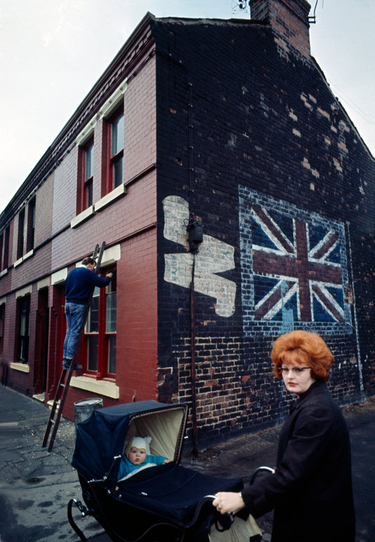 A photograph featuring a part of a building with a 'Union Jack' flag design on its side, with a man working on the building's facade and a woman holding with a baby stroller.