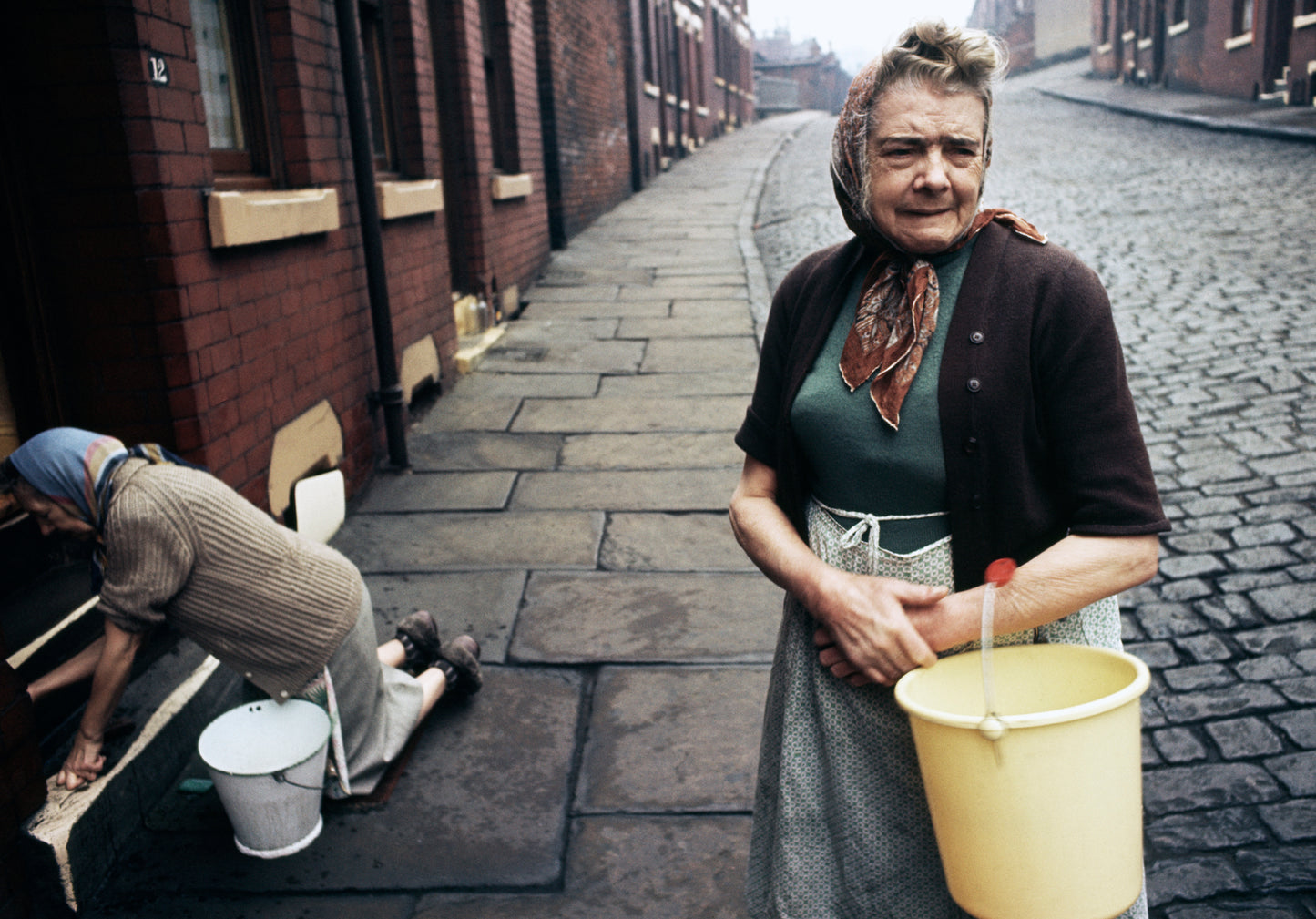 A photograph of an old lady holding a yellow bucket with another lady cleaning a staircase