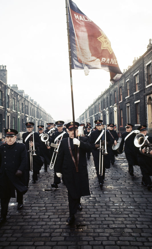 A photograph of a brass band marching down a street, with buildings on either side.