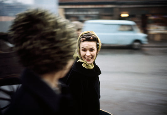 A photograph of a woman with a smile on her face, seen from behind, standing on a street corner.
