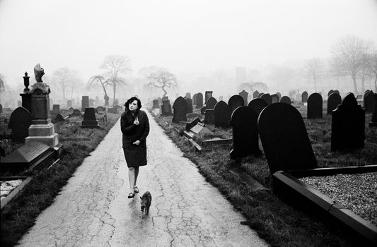 A black and white photograph of a woman at a cemetery accompanied by a cat