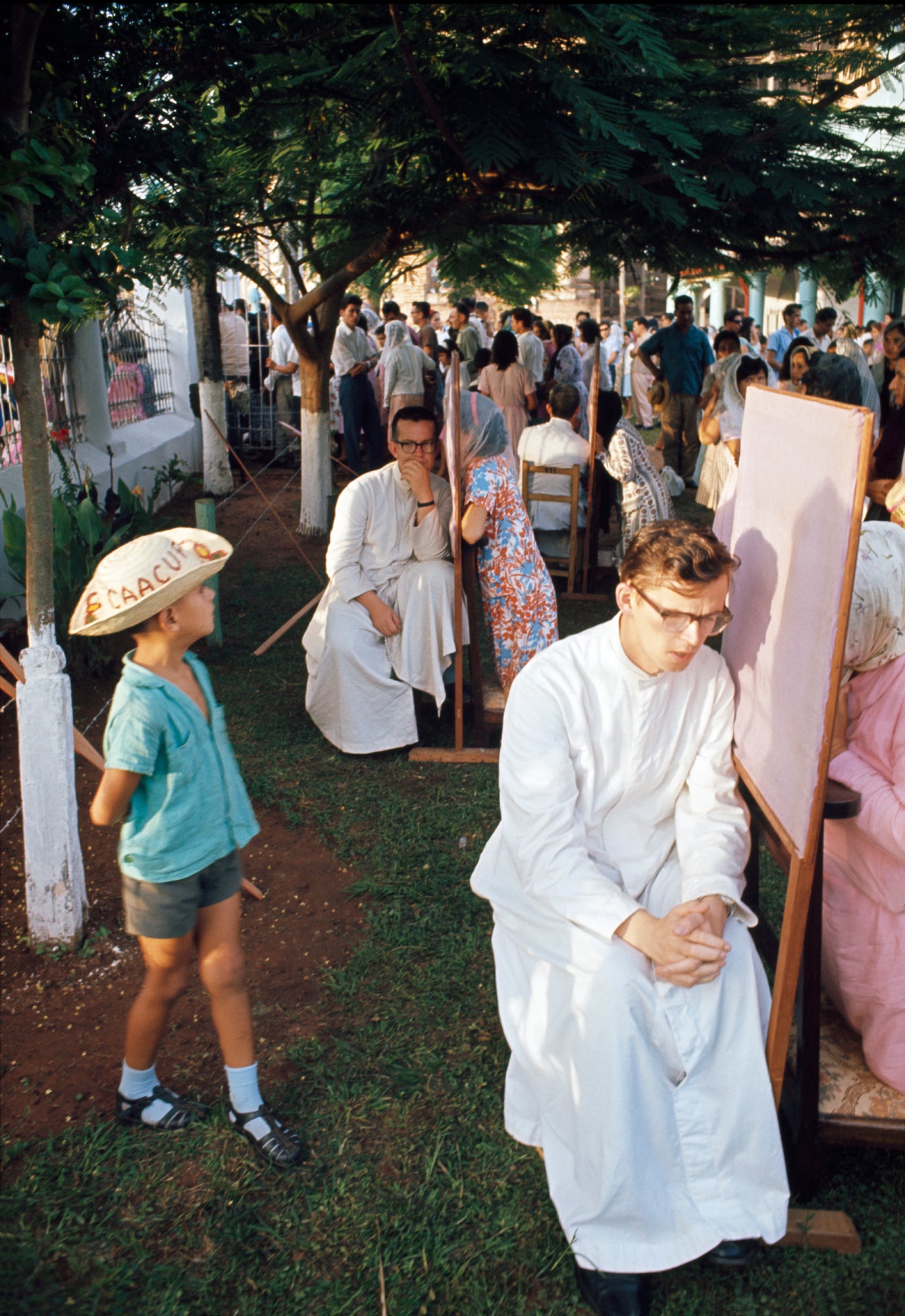 A photograph featuring a group of people seated and standing around a bulletin board, engaged in a gathering with priests, with a child looking on.