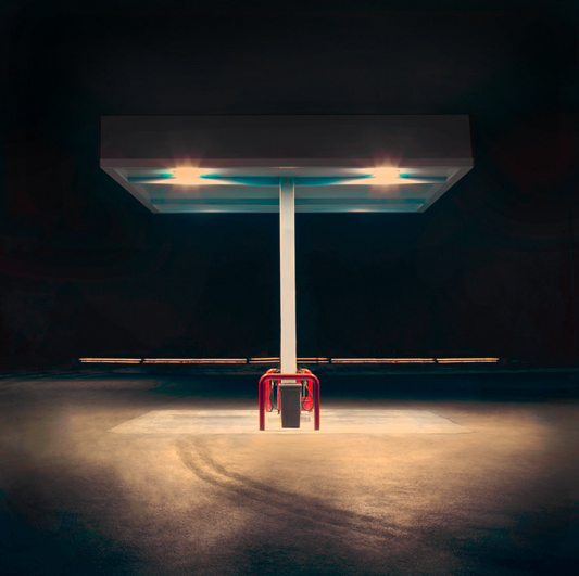A photograph featuring a petrol station with a white pump and a red canopy, illuminated by the headlights of a car.