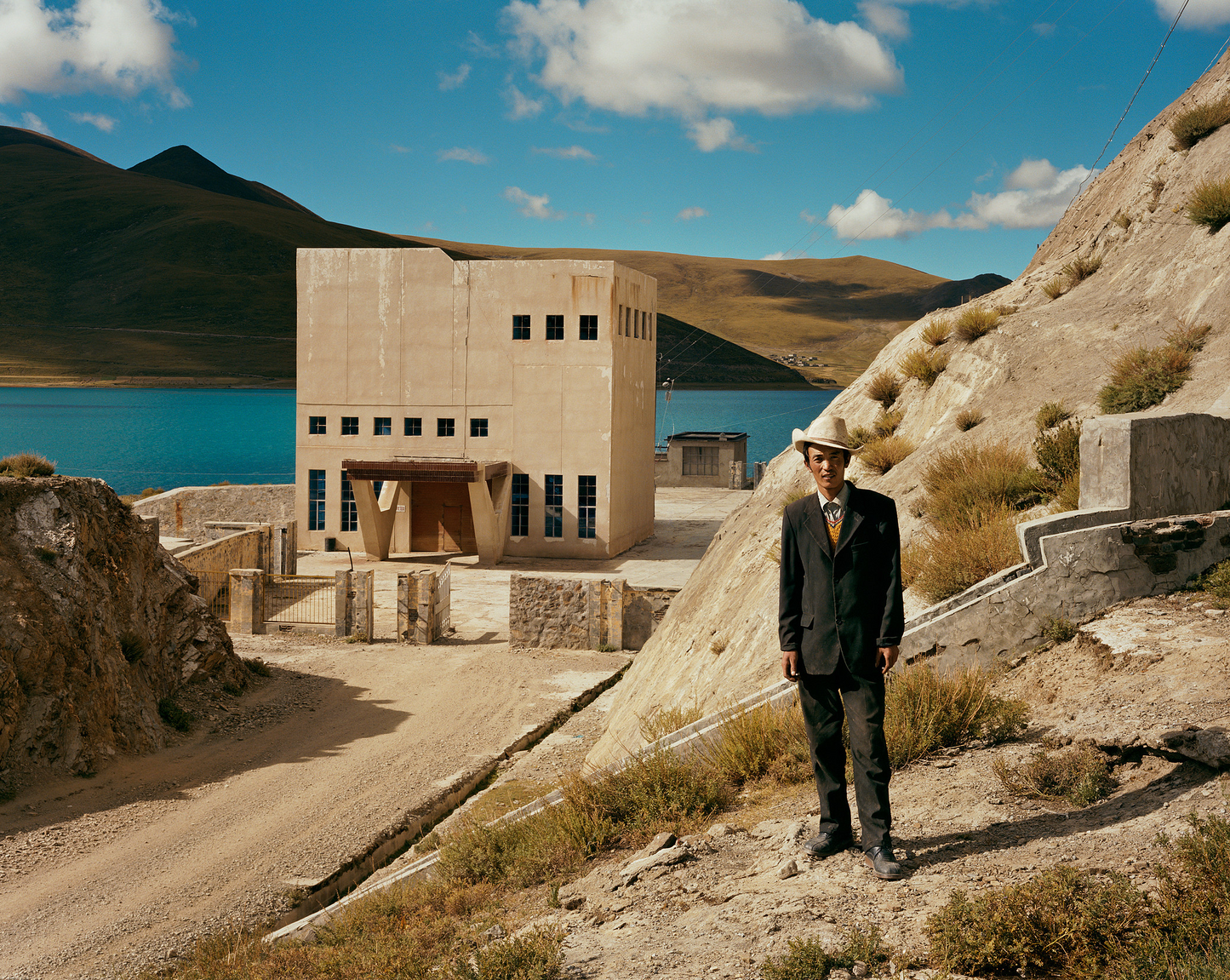 A man standing on a mountainous terrain with a large building in the background, which is part of a photographic print series by Barry Cawston - Tibetan Cowboy.