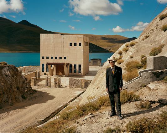 A man standing on a mountainous terrain with a large building in the background, which is part of a photographic print series by Barry Cawston - Tibetan Cowboy.