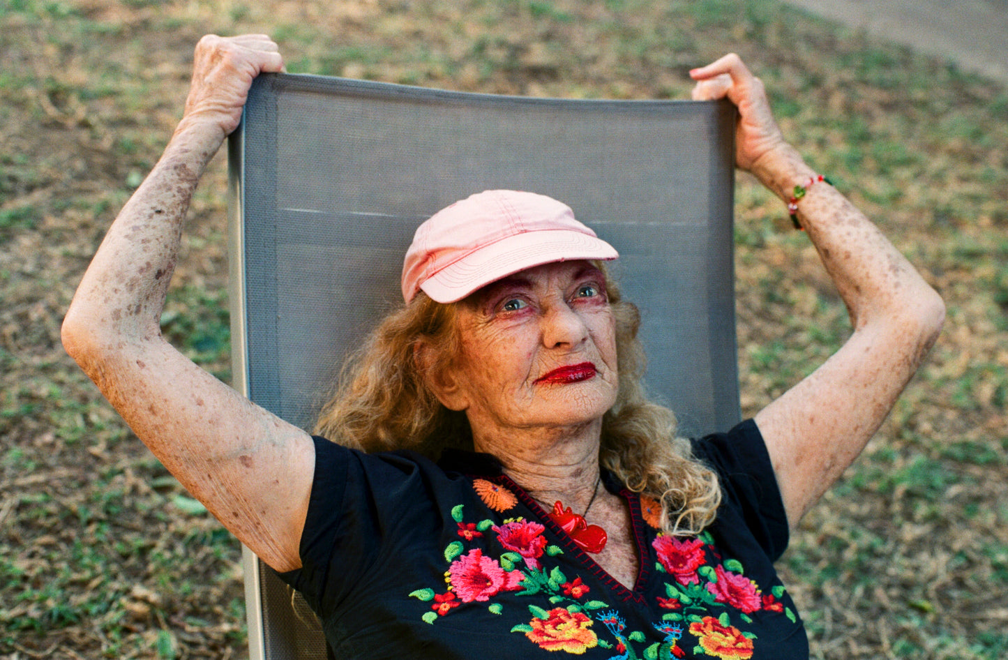 A photograph of an elderly woman with a pink hat and floral top, sitting in a chair outdoors.
