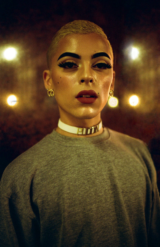 A close-up portrait of a person with a bold facial expression, wearing a grey t-shirt and a white choker necklace.