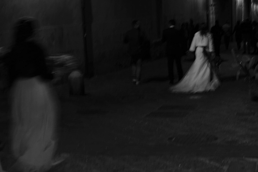 A black and white image of a group of people, likely a bride and groom, walking down a street at night, with the bride in a white dress and a tiara.