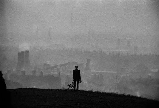 Black and white photograph of an industrial landscape with a figure standing on a hill with a dog overlooking the scene.