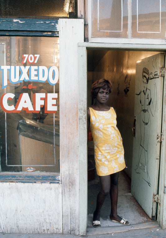 A photograph of a woman in a yellow dress standing in front of a sign that reads 'Tuxedo Cafe'.