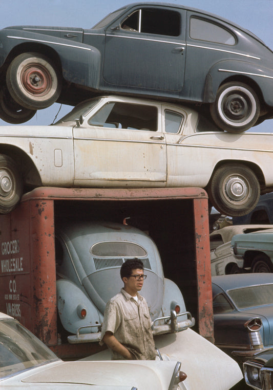 A photograph of a  mechanic standing in front of a stack of old cars