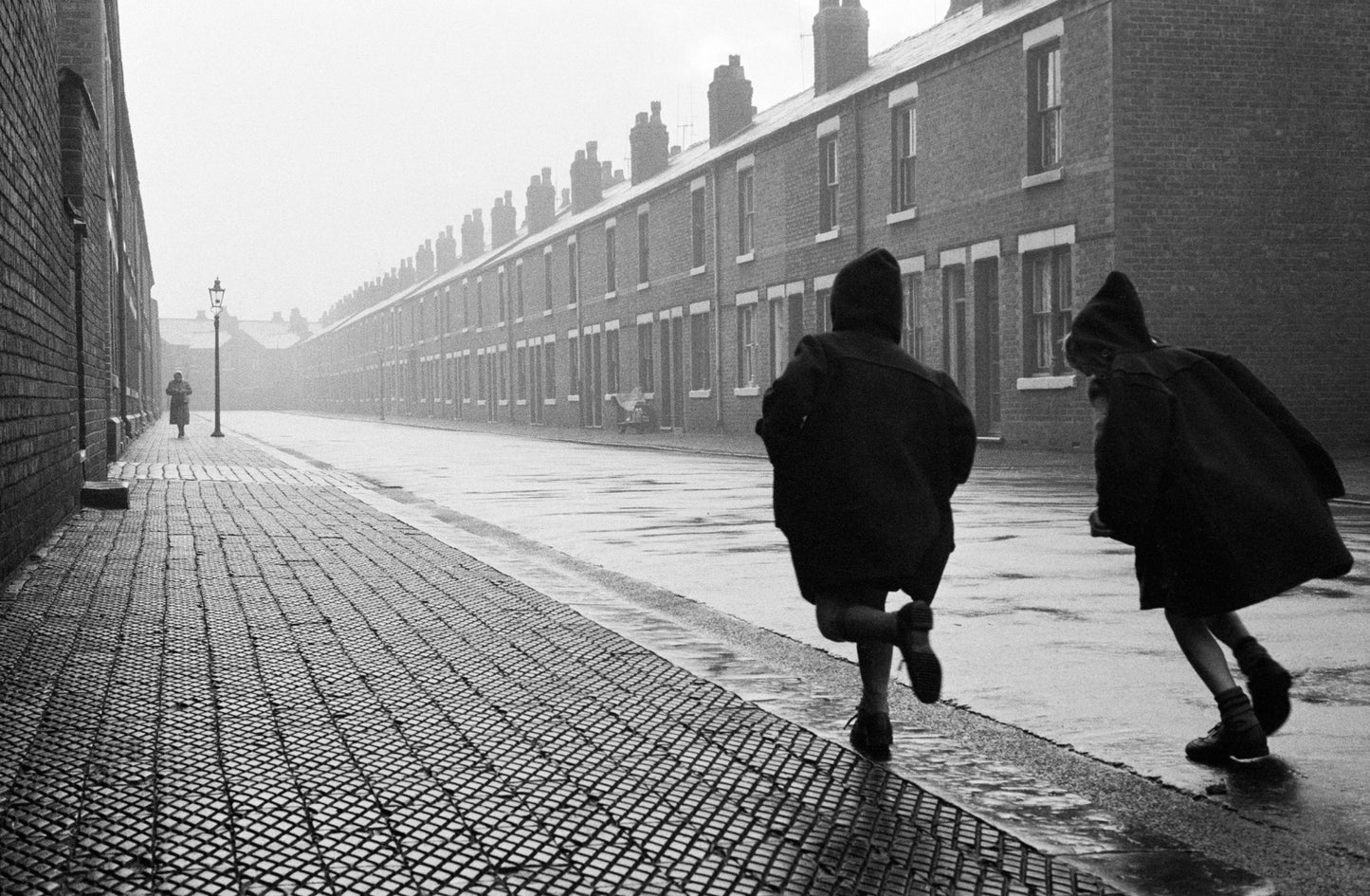 Black and white photograph of two children playing in a rain-soaked street, with buildings lining the road.