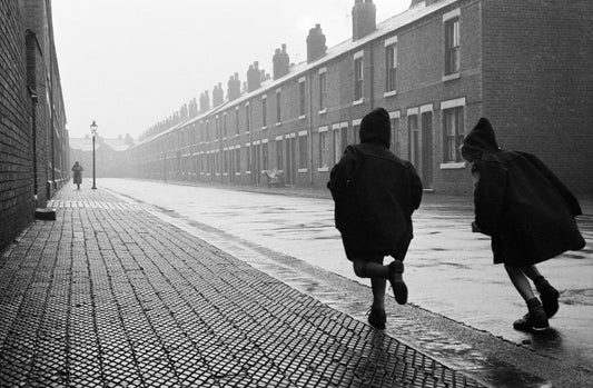 Black and white photograph of two children playing in a rain-soaked street, with buildings lining the road.