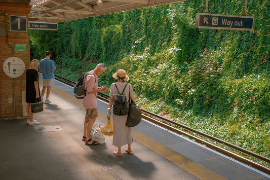 An image featuring a train station platform with two people standing on the platform, surrounded by greenery and train tracks.